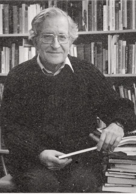 Black and white portrait of Noam Chomsky sitting in front of a wall of books.